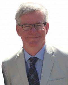 Head and shoulders photo of Martin Hendry. He is a white main, with short white hair, wearing glasses. He is wearing a blue shirt, dark blue tie and a cream jacket.